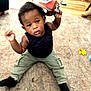 child, toddler, indoor, carpet, floor, cap, black_shirt, green_pants, socks, toy, wooden_floor, playful, cute, person, baby, sitting, curious, casual_clothing, home, young_child