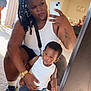 woman, child, toddler, mirror, selfie, braided_hair, indoor, living_room, carpet, smartphone, white_tank_top, denim_shorts, sneakers, gold_watch, tattoo, earrings, chandelier, vertical_blinds, casual, family