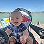 Marcelo is registered to the contest to win money with this photo: baby, child, smiling, happy, outdoor, lake, water, blue_sky, mountain, chair, fan, onesie, tie_dye, shark_design, sunny, nature, rock, playful, summer, cute