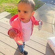 Jordyn is registered to the contest to win money with this photo: toddler, child, pink_jacket, snack_pouch, jeans, black_shoes, wooden_deck, sunlight, outdoor, grass, sidewalk, curious_expression, messy_hair, earring, daylight, casual_clothing, person, street, vehicle, shadow
