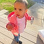 toddler, child, pink_jacket, snack_pouch, jeans, black_shoes, wooden_deck, sunlight, outdoor, grass, sidewalk, curious_expression, messy_hair, earring, daylight, casual_clothing, person, street, vehicle, shadow