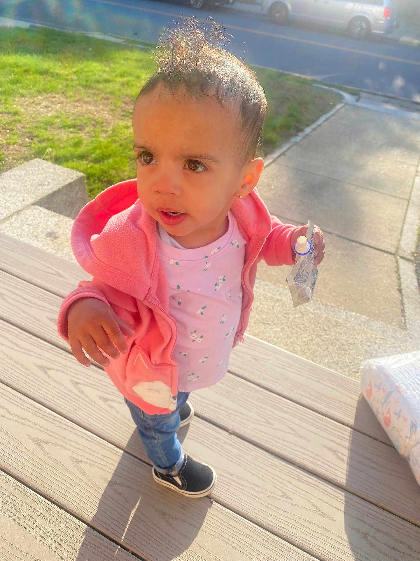 Jordyn is registered to the contest to win money with this photo: toddler, child, pink_jacket, snack_pouch, jeans, black_shoes, wooden_deck, sunlight, outdoor, grass, sidewalk, curious_expression, messy_hair, earring, daylight, casual_clothing, person, street, vehicle, shadow