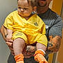 man, baby, child, tattoos, cap, yellow_gown, orange_socks, hospital, indoor, floor_tiles, holding, person, expression, casual_clothing, short_hair, beard, door, wall, clinic, hospital_wristband