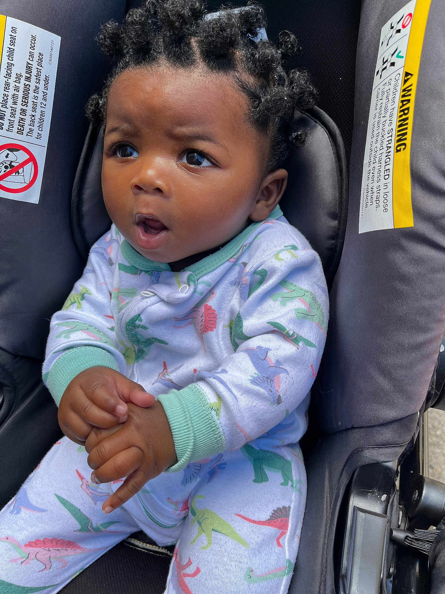 Kolby joined the competition — help win amazing prizes! baby, child, car_seat, dinosaur_pajamas, curly_hair, bantu_knots, hands, expression, wide_eyes, infant, seatbelt_warning_labels, clothing, portrait, cute, indoors, seat, person, young_child, sitting, closeup