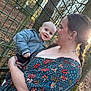 baby, woman, child, outdoor, smiling, holding, jacket, floral_dress, fence, trees, sunlight, nature, forest, happy, portrait, person, braid, tattoo, fall_leaves, daylight