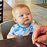 Grayson joined the competition — help win amazing prizes! baby, child, high_chair, restaurant, indoor, face, portrait, serious_expression, pout, blue_eyes, blond_hair, short_hair, toddler, snack, food, adult_hand, table, wooden_table, nike_shirt, dining_area