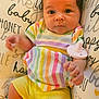 baby, infant, child, colorful_clothing, striped_shirt, yellow_shorts, pacifier, pillow, text_print, script_text, cute, curious_eyes, soft_lighting, indoor, portrait, close_up, human, skin, hand, face