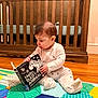 baby, infant, crib, book, board_book, play_mat, pajamas, hardwood_floor, nursery, child, sitting, reading, zebra, colorful, indoor, portrait, floor, toy, furniture, curious