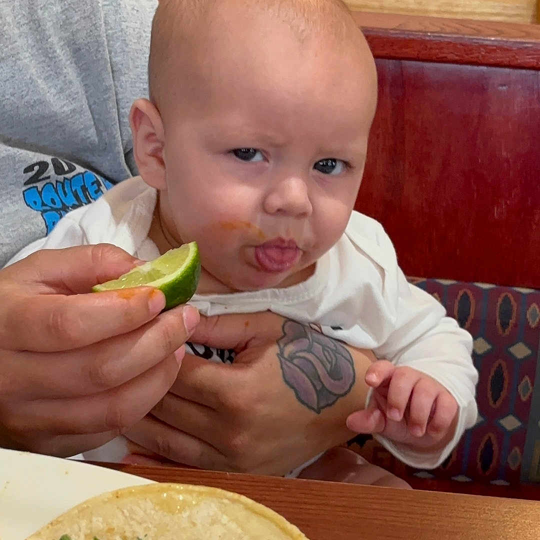 Bennett is registered to the contest to win money with this photo: baby, child, food, lime, taco, restaurant, table, hand, tattoo, face, expression, sour, sitting, clothing, person, plate, indoor, furniture, booth, meal