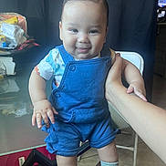 Matthew is registered to the contest to win money with this photo: baby, child, smiling, blue_overalls, standing, hand_support, indoor, table, clutter, chair, socks, shoes, face, person, cute, infant, happy, portrait, skin, hair