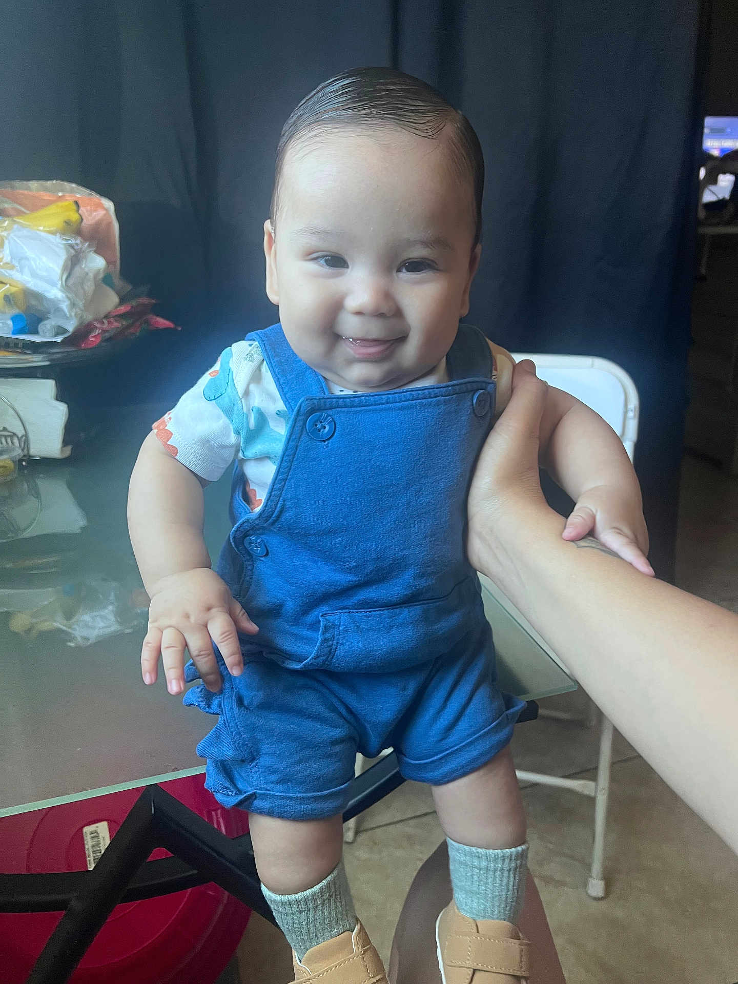 Matthew is registered to the contest to win money with this photo: baby, child, smiling, blue_overalls, standing, hand_support, indoor, table, clutter, chair, socks, shoes, face, person, cute, infant, happy, portrait, skin, hair