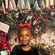 Deomi is registered to the contest to win money with this photo: child, girl, smiling, red_bow, black_dress, christmas, holiday, decorations, candles, pine_branches, berries, gift, stocking, festive, indoors, portrait, happy, celebration, seasonal, background