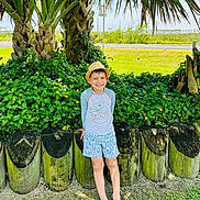 Erick joined the competition — help win amazing prizes! child, boy, smile, hat, palm_tree, tropical_plants, yellow_flowers, wooden_posts, grass, sandals, shorts, rash_guard, standing, outdoors, sunny, hedge, tree_trunk, greenery, portrait, vacation