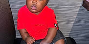 Lennox is registered to the contest to win money with this photo: toddler, child, sitting, red_shirt, black_shorts, sneakers, white_shoes, black_shoes, curly_hair, indoor, bench, cushion, modern_wall, wooden_ledge, flash_reflection, looking_at_camera, cute, portrait, young_child, casual_clothing