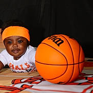 Lennox is registered to the contest to win money with this photo: toddler, child, basketball, orange_headband, white_shirt, sports, indoor, toy, playing, cute, baby, face, smiling, person, ball, reaching, activity, black_background, floor, colorful