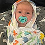 baby, pacifier, blanket, dinosaur_pattern, wrapped, infant, face, wide_eyes, cute, child, soft_fabric, colorful, pattern, indoors, portrait, head, expression, comfort, seated, clothing