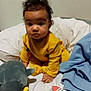 baby, child, yellow_outfit, curly_hair, bed, blanket, toy, stuffed_animal, sock, blue_blanket, indoor, person, infant, cute, expression, soft_toys, colorful, playtime, portrait, sitting