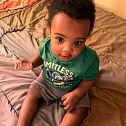 Kantrell joined the competition — help win amazing prizes! baby, child, infant, bed, blanket, green_shirt, shorts, indoors, person, cute, sitting, looking_up, skin, hair, face, expression, young_child, comfortable, cozy, casual_clothing