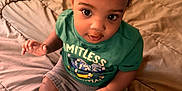 Kantrell is registered to the contest to win money with this photo: baby, child, infant, bed, blanket, green_shirt, shorts, indoors, person, cute, sitting, looking_up, skin, hair, face, expression, young_child, comfortable, cozy, casual_clothing