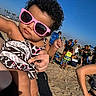 baby, child, sunglasses, beach, sand, summer, people, sunny, patterned_clothing, curly_hair, outdoor, vacation, family, person, holding, footwear_barefoot, blue_sky, casual, fun, relaxation