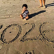 Amira is registered to the contest to win money with this photo: baby, beach, sand, footprints, year, 2026, child, curious, sunny, shadow, legs, sitting, outdoor, daytime, barefoot, casual, nature, summer, holiday, playful