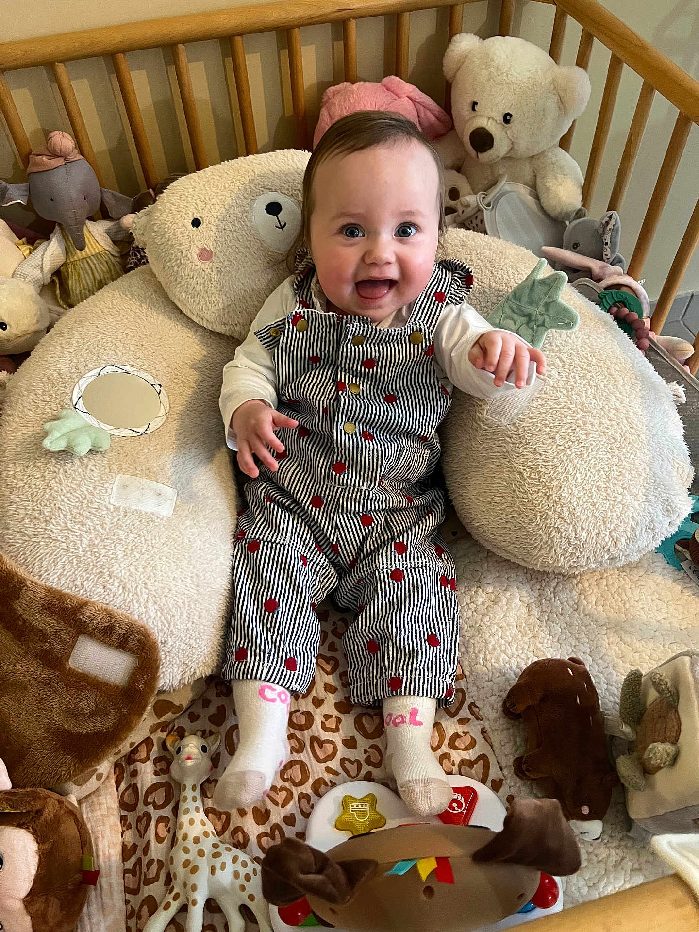 Luna a rejoint le concours — aidez-le/la à gagner de superbes lots ! baby, infant, smiling, crib, stuffed_animal, teddy_bear, giraffe_toy, plush_pillow, striped_overalls, socks, play_mat, toy, nursery, indoor, portrait, happy, seated, camera_eye_contact, soft_toys, boppy_pillow