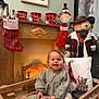 Louise a rejoint le concours — aidez-le/la à gagner de superbes lots ! toddler, child, smile, plush_toy, wooden_cradle, christmas_stocking, fireplace, holiday_decor, teddy_bear, festive, indoor, clothing, pillow, hat, lantern, painting, holiday_candle, cozy, happy, winter