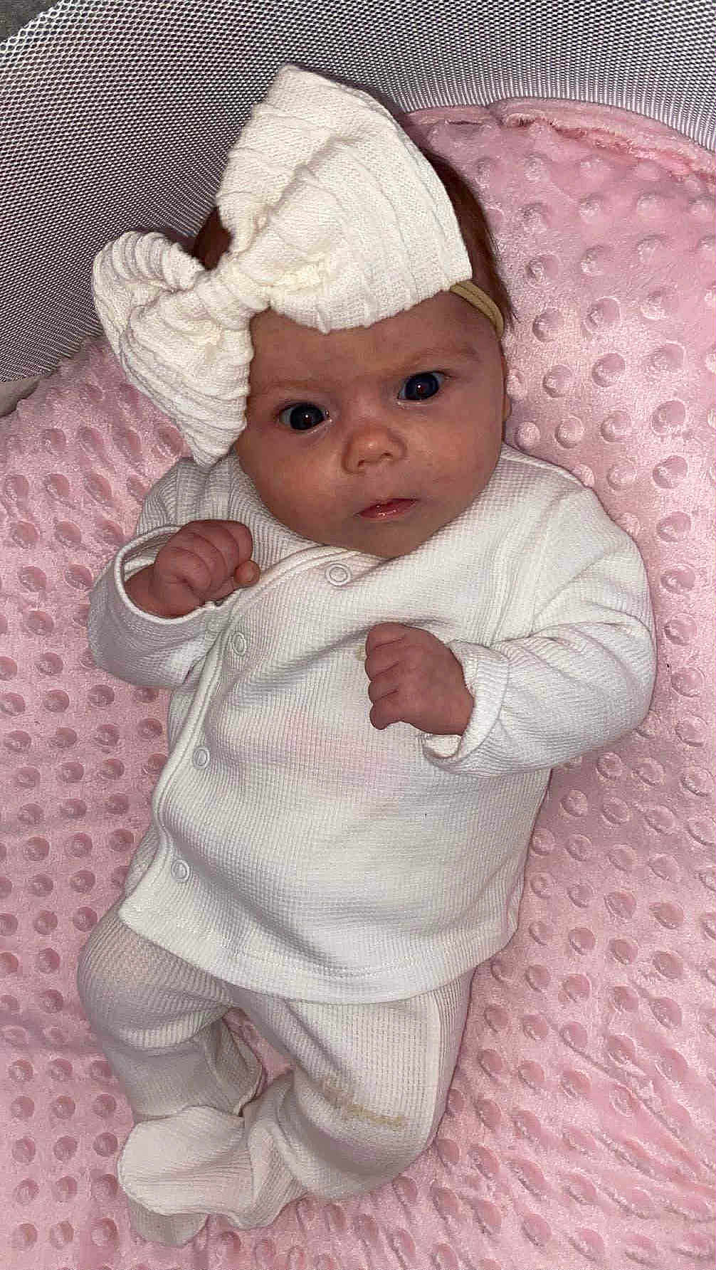 Ella joined the competition — help win amazing prizes! baby, infant, child, white_clothing, headband, bow, pink_blanket, texture, soft, cute, lying_down, portrait, indoors, newborn, small_hands, expression, cozy, sleepy, face, person