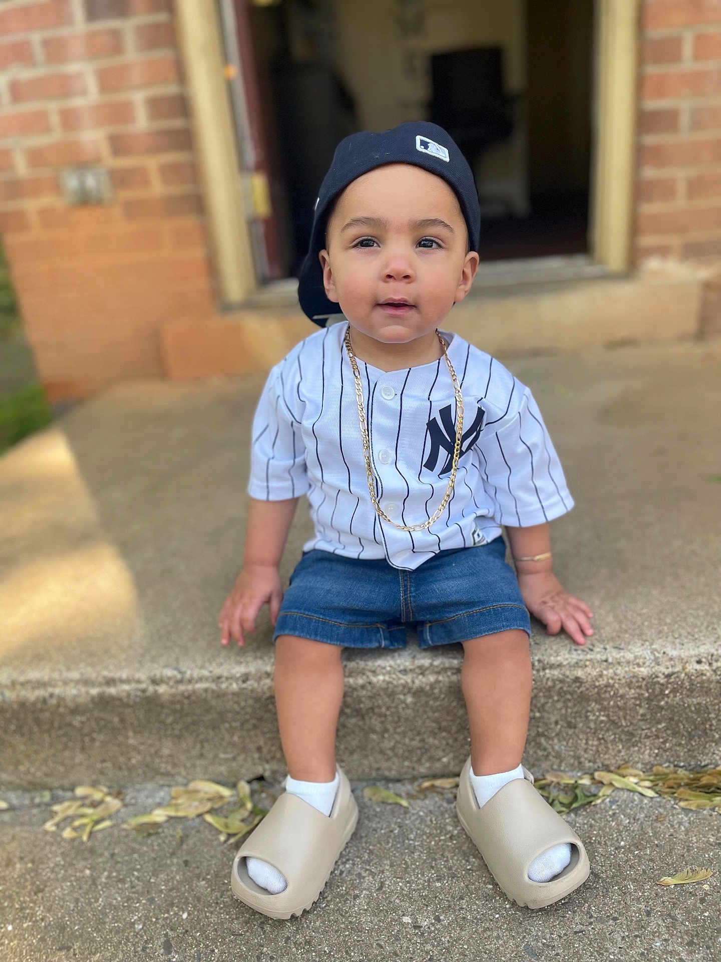 Jacob joined the competition — help win amazing prizes! toddler, child, sitting, baseball_cap, white_jersey, denim_shorts, gold_chain, concrete_step, outdoor, footwear, socks, casual_clothing, curious_expression, person, portrait, daylight, sidewalk, brick_wall, street_style, young_child