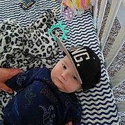 Adriel joined the competition — help win amazing prizes! baby, cap, blanket, plush_toy, remote_control, patterned_bedding, leopard_print, cradle, carpet, hands, child, indoor, infant, clothing, person, blue, black, white, face, toy
