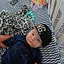 baby, cap, blanket, plush_toy, remote_control, patterned_bedding, leopard_print, cradle, carpet, hands, child, indoor, infant, clothing, person, blue, black, white, face, toy