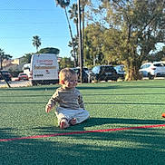 Valentin a rejoint le concours — aidez-le/la à gagner de superbes lots ! toddler, child, outdoor, sports_court, barefoot, sunlight, green_surface, trees, cars, fence, smile, sitting, casual_clothing, daytime, playground, young_child, recreation, park, nature, leisure