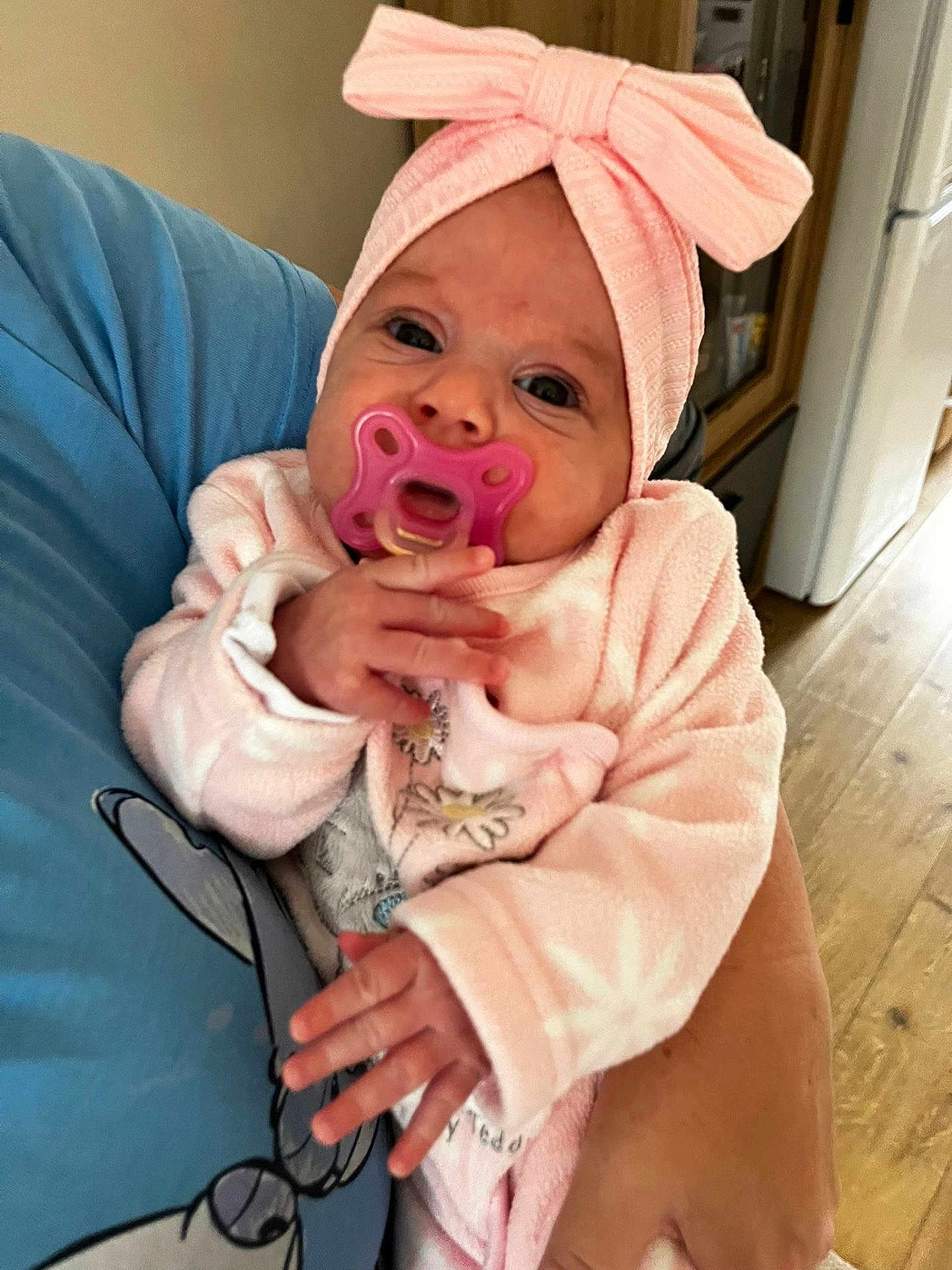 Lilibeth joined the competition — help win amazing prizes! baby, infant, pacifier, pink_clothing, headband, bow, hand, adult_hand, blue_shirt, cartoon_character, indoor, wooden_floor, cabinet, cozy, closeup, portrait, person, cute, newborn, holding