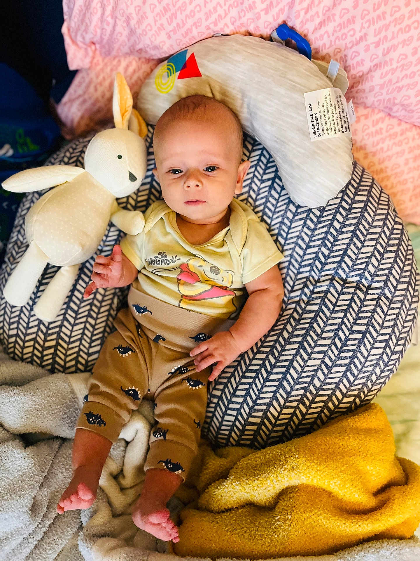 Noah is registered to the contest to win money with this photo: baby, infant, stuffed_toy, rabbit_toy, pillow, blanket, cozy, clothing, patterned_pillow, yellow_blanket, soft_toy, child, indoors, cute, relaxed, lying_down, person, foot, hand, face