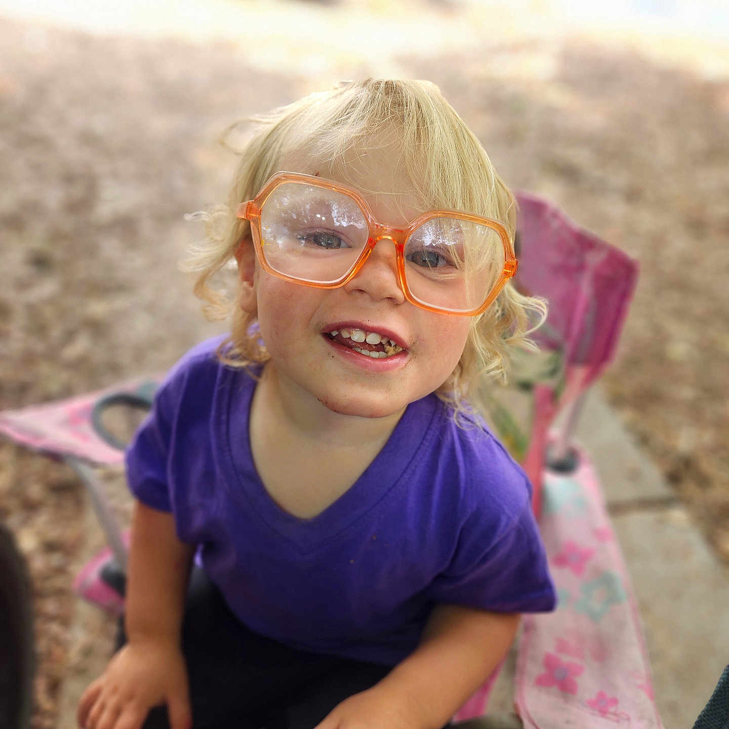 Thorin is registered to the contest to win money with this photo: blonde_hair, child, closeup, curly_hair, cute, daylight, face, glasses, happy, nature, orange_glasses, outdoor, person, pink_chair, playful, purple_shirt, sitting, smiling, toddler, young