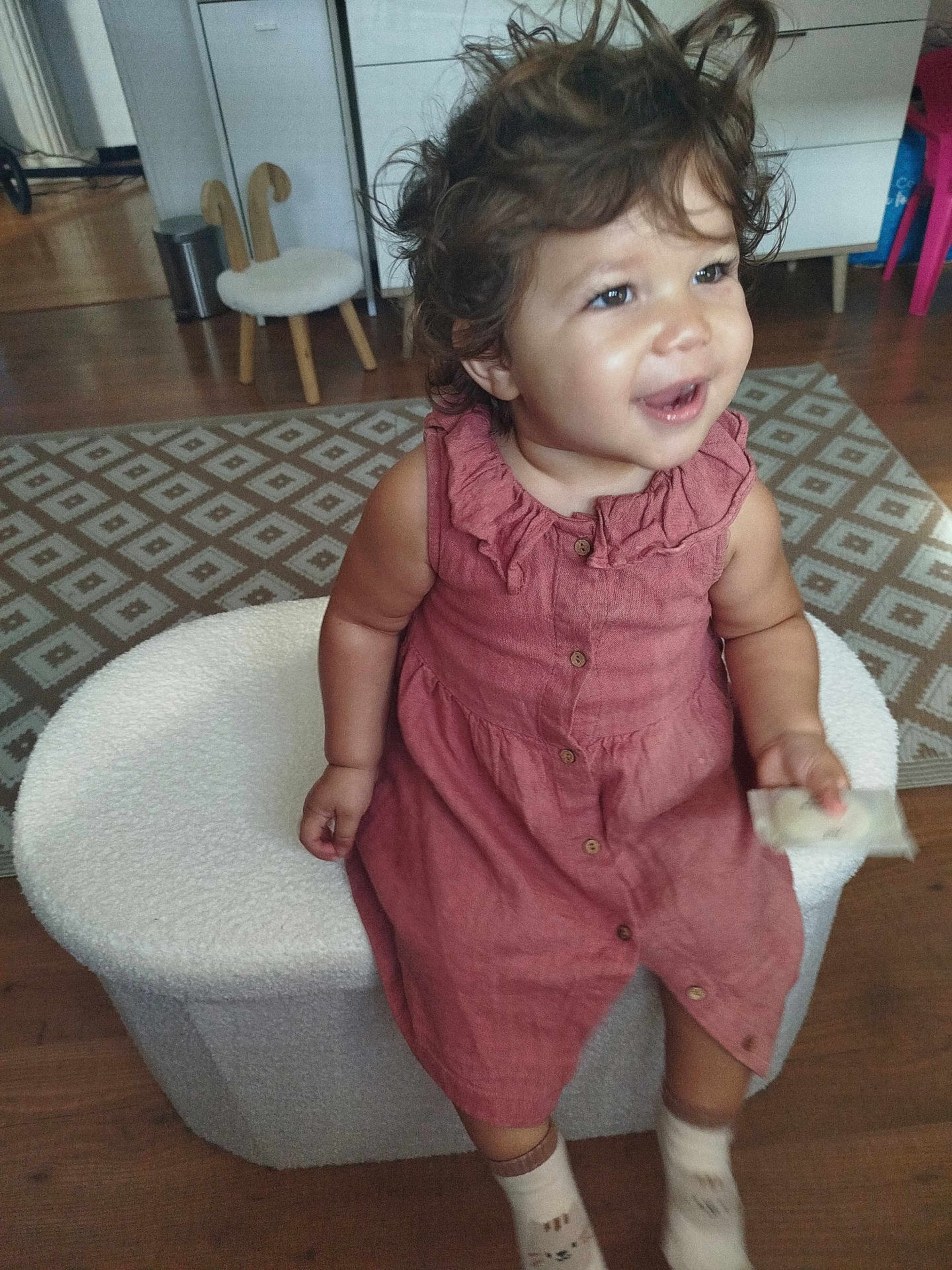 Ohana participe au concours pour gagner de l'argent avec cette photo : toddler, child, curly_hair, dress, pink_dress, socks, smiling, indoors, ottoman, rug, wooden_floor, furniture, chair, toy, happy, person, cute, home, small_object, playful