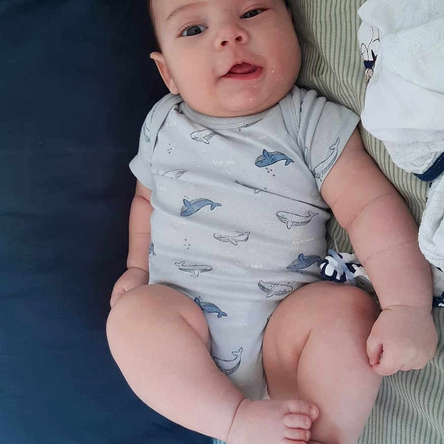 Benicio a rejoint le concours — aidez-le/la à gagner de superbes lots ! baby, infant, onesie, whale_pattern, pillow, bed, blanket, drool, cute, chubby_cheeks, skin, lying_down, striped_sheet, blue_pillow, white_blanket, sleepwear, smiling, human, child, indoors