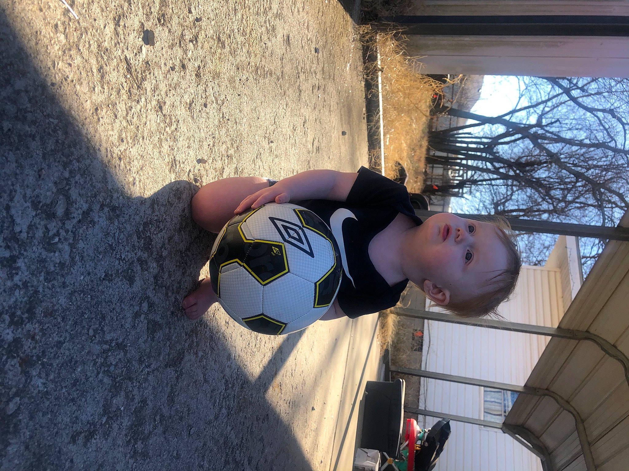 Aiden is registered to the contest to win money with this photo: automotive_exterior, ball, child, concrete, eyewear, football, fun, human_leg, leisure, person, personal_protective_equipment, plant, reflection, shadow, tints_and_shades, toddler, toy, tree, vehicle_door, window