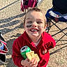 bread, candid, casual_clothing, child, daytime, face_paint, fence, folding_chair, grass, green_soda_can, happy, outdoor, person, playful, portrait, red_jacket, seated, smiling, sunlight, young_child