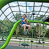 active, blue_shirt, child, climbing, daylight, fun, green, happy, nature, outdoor, park, playground, recreation, rope_structure, shorts, sky, smiling, sneakers, trees, young