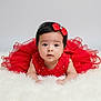 baby, child, red_dress, bow, cute, portrait, infant, fluffy, white_background, face, adorable, fashion, tutu, lying_down, young_child, studio_photo, black_hair, looking_at_camera, decorative_clothing, soft_texture