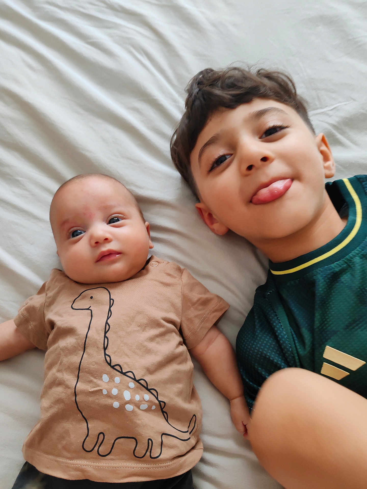 Faël participe au concours pour gagner de l'argent avec cette photo : baby, child, dinosaur_shirt, bed, playful, tongue_out, cute, siblings, laying_down, casual_clothing, indoor, portrait, smile, happy, infant, young_child, family, person, bed_sheet, relaxed