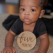 Gianni joined the competition — help win amazing prizes! baby, infant, five_months, carpet, wooden_sign, drool, black_shirt, curly_hair, close_up, face, hands, cute, indoors, child, portrait, young_baby, expression, skin, person, lying_down