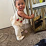 baby, child, standing, smiling, indoor, carpet, furniture, metal_shelf, decor, top_knot, clothing, cute, happy, person, home, floor, toy, pacifier, strawberry_pattern, curly_hair
