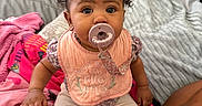 Xaila joined the competition — help win amazing prizes! baby, pacifier, pink_bib, curly_hair, bed, blanket, pillow, child, indoor, person, cute, sitting, clothing, hand, footwear, furniture, room, soft_toys, colorful, expression