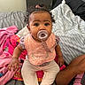 baby, pacifier, pink_bib, curly_hair, bed, blanket, pillow, child, indoor, person, cute, sitting, clothing, hand, footwear, furniture, room, soft_toys, colorful, expression
