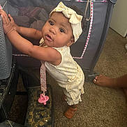 Xaila is registered to the contest to win money with this photo: baby, child, standing, headband, yellow_outfit, indoor, carpet, playpen, toy_pacifier, footwear, curly_hair, furniture, hand, expression, person, infant, home, floor, looking_up, balance