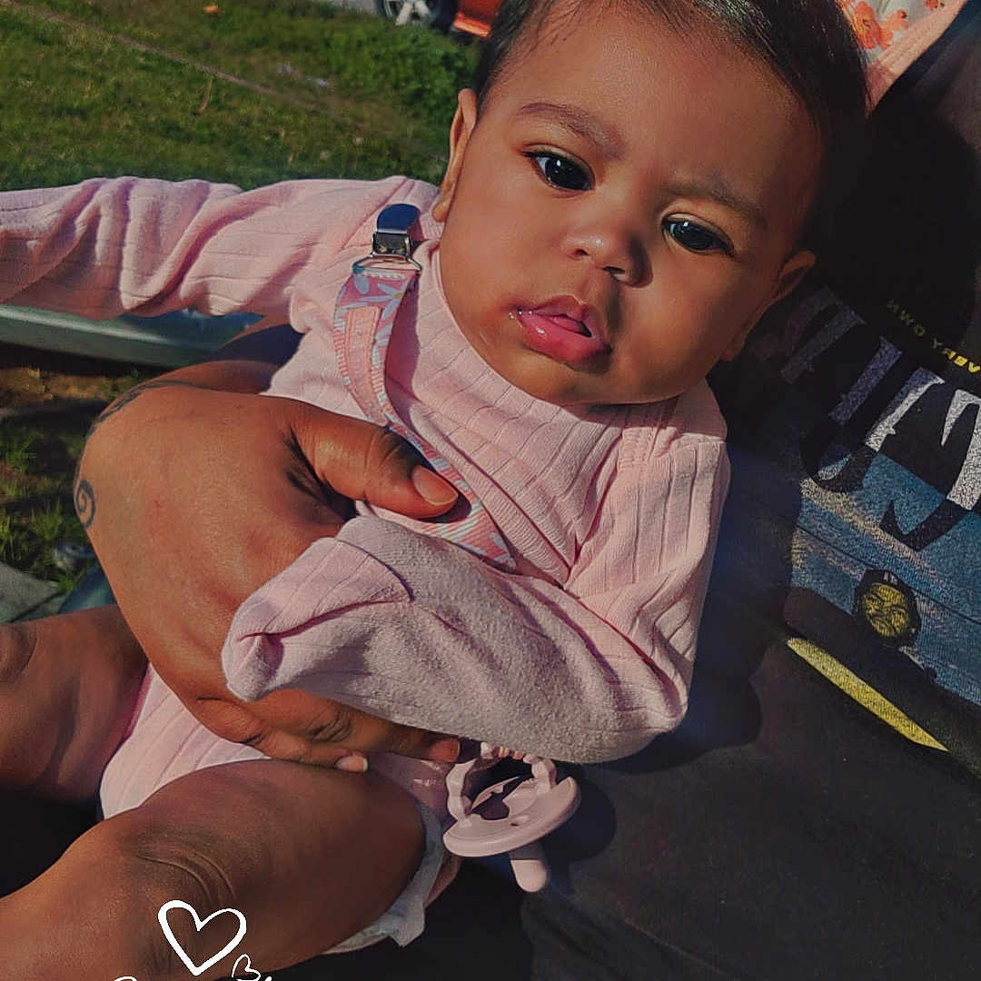 Xaila is registered to the contest to win money with this photo: baby, child, infant, holding, pink_clothing, outdoor, daylight, grass, car, suburban, person, hand, blanket, pacifier, face, expression, portrait, sunlight, nature, casual