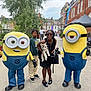 dog, person, child, mascot, costume, minion, outdoor, street, urban, building, pavement, tree, sunglasses, black_clothing, casual, smile, festival, colorful, handbag, footwear