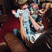 Jason joined the competition — help win amazing prizes! baby, cap, adult, living_room, couch, carpet, blanket, person, smile, furniture, seated, indoor, hands, plaid_blanket, curtains, jeans, shirt, red_carpet, wood_paneling, happy